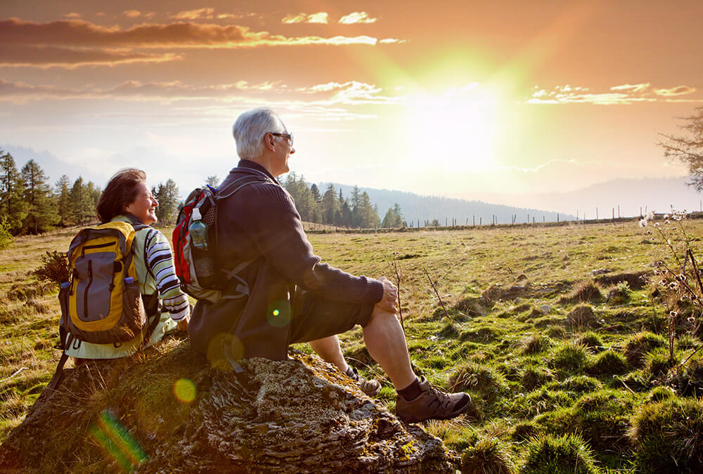 Two older adults with backpacks sit on a rock in a grassy field, admiring a scenic sunset over distant trees and hills, enjoying the outdoors and nature together.